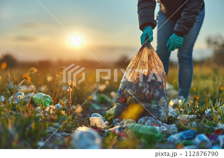 AI-generated content. Volunteer in gloves stands holding a bag of litter in a field, illuminated by the soft light of dawn. AI-generated content. Volunteer in gloves stands holding a bag of litter in a field, illuminated by the soft light of dawn. 112876790