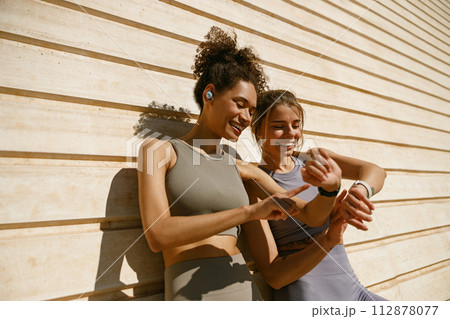 Smiling woman with female friend looking on smartwatch before exercising standing near wall outdoors 112878077