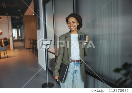 A woman, in formal wear, stands in a hallway with a cup of coffee and laptop 112878983