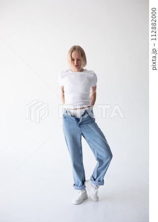 Beautiful blonde girl in a white T-shirt and blue jeans and sneakers posing on a white background 112882000