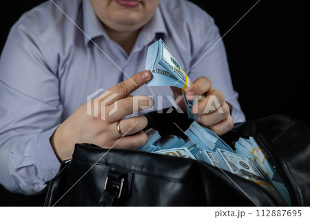 A pack of dollars in the hands of a man on a black background close-up. A rich man sits over a bag full of money and counts stacks of hundred dollar bills. 112887695