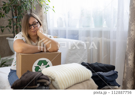 A woman is thoughtful with a box with a recycling sign in her hands while sorting out clothes. The woman was thinking about recycling her clothes or donating them to a charity. A woman is thoughtful with a box with a recycling sign in her hands while sorting out clothes. The woman was thinking about recycling her clothes or donating them to a charity. 112887756