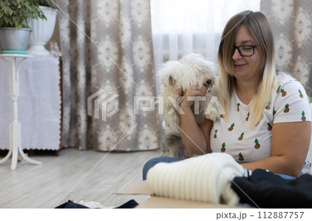 While a woman sorts through her clothes and puts them in a donation box, a small dog plays nearby. The dog prevents the woman from sorting through her clothes by playing with her. While a woman sorts through her clothes and puts them in a donation box, a small dog plays nearby. The dog prevents the woman from sorting through her clothes by playing with her. 112887757
