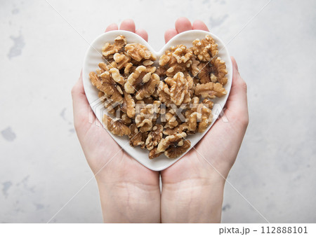 Female hands holding heart shaped plate with healthy peeled walnut nuts on light background. 112888101
