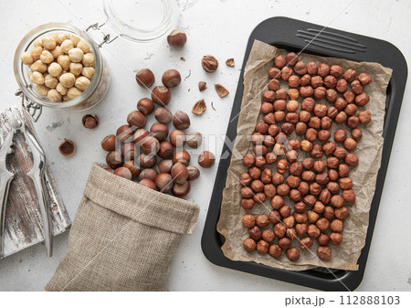 Baking tray with raw hazelnut nuts with cracker and peeled nuts in jar on light kitchen background. 112888103