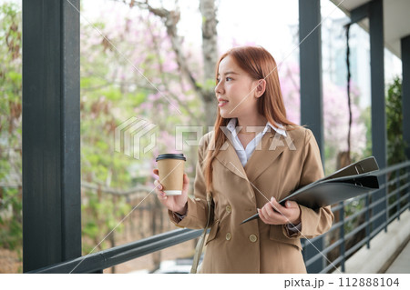 A young Asian businesswoman wearing a suit holding a cup of coffee and files walking in a big city. Young Asian businesswomen walking to the office. A young Asian businesswoman wearing a suit holding a cup of coffee and files walking in a big city. Young Asian businesswomen walking to the office. 112888104
