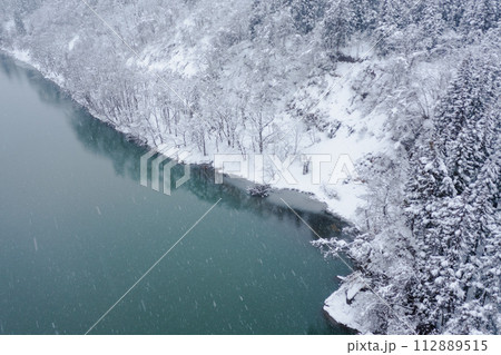 ドローン写真：雪降る冬、雪に覆われた真っ白な木々とエメラルドグリーンの川	 112889515