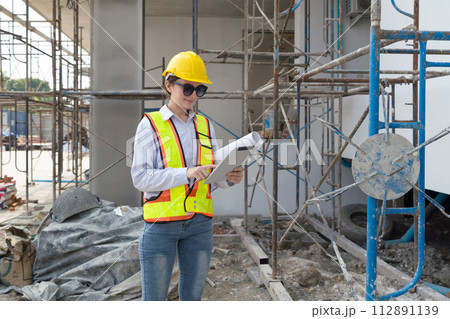 Young female engineer in a construction helmet with a floor plan stands by metal scaffolding, typing on tablet computer. Young female engineer in a construction helmet with a floor plan stands by metal scaffolding, typing on tablet computer. 112891139