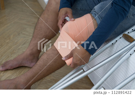 A man with a broken knee sitting on his bed. He leans on crutches, signalling recovery from injury. His bedroom serves as a backdrop, tinged with hints of helplessness yet enduring determination. 112891422