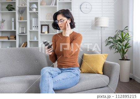Adult female sits on the sofa at home, reading a message on her smartphone, appearing confused and concerned after receiving bad news. 112895215
