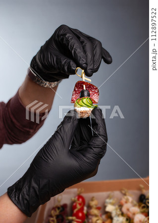 Canape, hands in black gloves hold a beautiful canape on a cap. Catering, snacking, food. Studio photo. 112897522