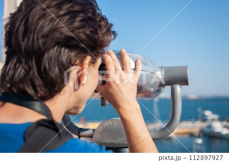 man looks through stationary binoculars. Binoscope on observation deck near sea. seaport in Odessa. Expensive yachts on the pier. Commercial machine, vending business. 112897927