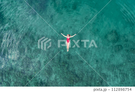 Aerial View of a Woman in Red Swimsuit Floating Serenely on the Crystal Clear Ocean Waters During Midday 112898754