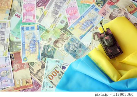 Toy tank on Ukrainian flag on many banknotes of different currency. Background of war funding and military support price in Ukraine 112903366