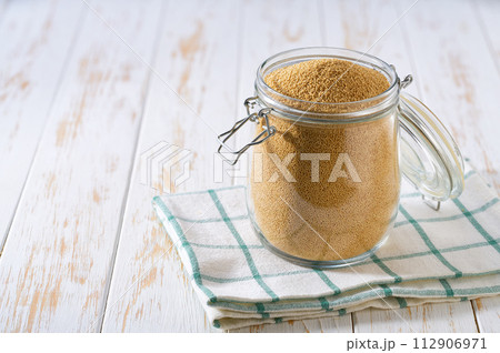Uncooked dry amaranth grains in glass storage jar on a white wooden table. 112906971