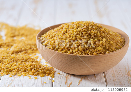 Raw bulgur are scattered out of the wooden bowl on a light table, selective focus. 112906982