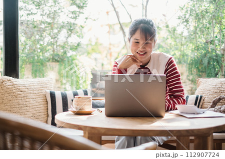 Cheerful woman enjoys a productive day working on her laptop in a well lit, plant-filled cafe, exuding a sense of satisfaction and comfort with a warm beverage by her side. Cheerful woman enjoys a productive day working on her laptop in a well lit, plant-filled cafe, exuding a sense of satisfaction and comfort with a warm beverage by her side. 112907724