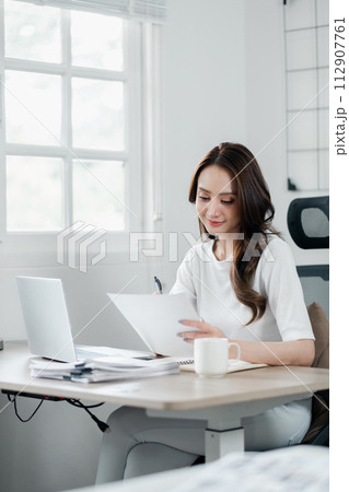 Businesswoman attentively reviewing documents, with a cup of coffee and laptop in her bright, airy home office. 112907761