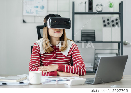 Businesswoman wearing a virtual reality headset sits confidently at her office desk, immersed in a digital workspace. 112907766