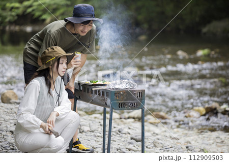 河原で焼き加減を真剣に見つめるバーベキューをする若い男女 河原で焼き加減を真剣に見つめるバーベキューをする若い男女 112909503