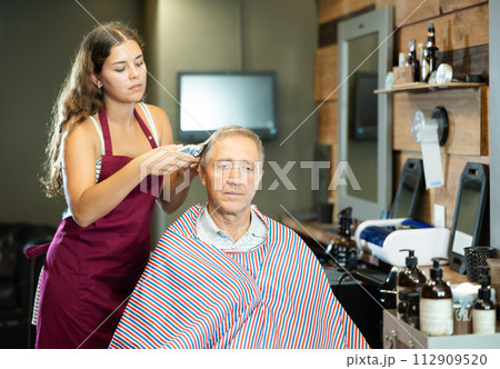 Young woman hairdresser performs haircut for elderly man using hair clipper 112909520