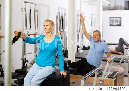 Mature couple using bed of Pilates reformer bed equipment while training together in wellness center. Wellbeing and healthy lifestyle concept 112909592