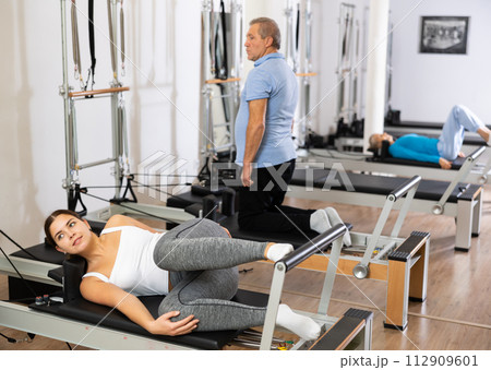 Focused young woman in activewear performing Pilates exercise for legs and back, training on bed equipment in gym 112909601