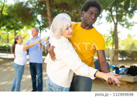 Exited older and middle-aged diverse people enjoying latin dances breaking into pairs on sandy area between trees 112909696