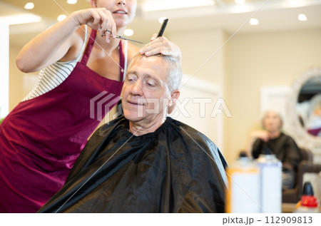 Young female hairdresser working with scissors and comb with mature man 112909813