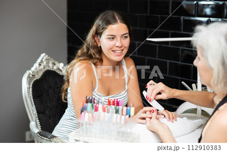 Friendly elderly woman working in beauty salon as manicure master shaping the nails of young female client with nail file Friendly elderly woman working in beauty salon as manicure master shaping the nails of young female client with nail file 112910383
