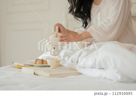 A woman in long dress pajamas pouring tea from a beautiful ceramic teapot, having breakfast in bed 112910895