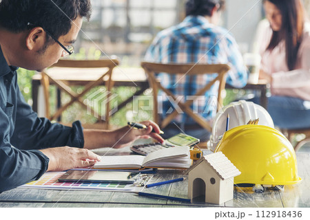 Civil construction engineer working with laptop at desk office with white yellow safety hard hat at office on construction site. Asian young man architecture project manager sitting at office on site 112918436