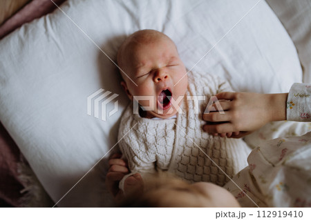 Top view of cute little baby yawning in bed, big sister holding newborn hand. Sisterly love, joy for new family member. 112919410