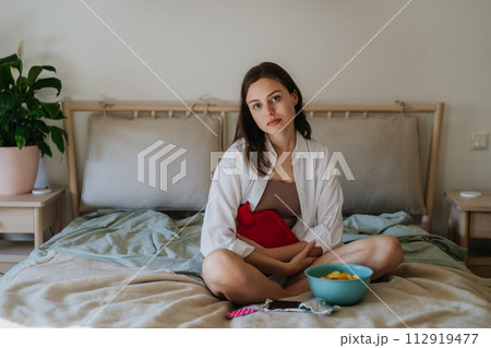 Woman at home suffering from menstrual pain, having cramps. Woman warming lower abdomen with a hot water bottle, endometriosis, and conditions causing pain in tummy. 112919477