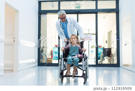 Friendly pediatrician pushing little patient in wheelchair. Cute preschool girl trusting her healthcare workers in hospital. 112919509