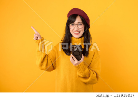 Portrait young beautiful 30s asian woman happy smile wearing yellow shirt and red hat pointing finger gesture to free copy space and using smartphone against yellow background. new application Portrait young beautiful 30s asian woman happy smile wearing yellow shirt and red hat pointing finger gesture to free copy space and using smartphone against yellow background. new application 112919817