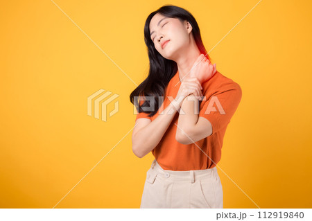 Attentive young Asian woman in her 30s, wearing an orange shirt, holds her painful neck on yellow background. office syndrome health care concept. Attentive young Asian woman in her 30s, wearing an orange shirt, holds her painful neck on yellow background. office syndrome health care concept. 112919840