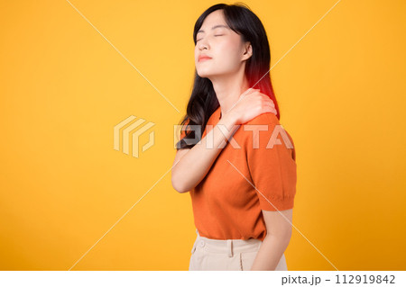 Neck ache therapy medical office syndrome concept. Concerned young Asian woman in her 30s, wearing an orange shirt, holds her pain shoulder on yellow background. Neck ache therapy medical office syndrome concept. Concerned young Asian woman in her 30s, wearing an orange shirt, holds her pain shoulder on yellow background. 112919842