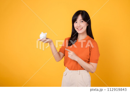 Smiling Asian woman in her 30s holding piggy bank, pointing to it, on vibrant yellow background. Wealth and saving concept. Smiling Asian woman in her 30s holding piggy bank, pointing to it, on vibrant yellow background. Wealth and saving concept. 112919843