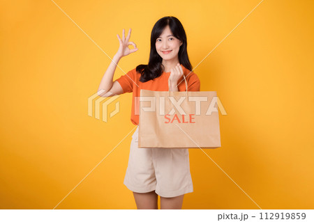 Celebrate savings and style in a shopping spree. Young woman holding a bag isolated on yellow background, radiating happiness in a mall setting. 112919859