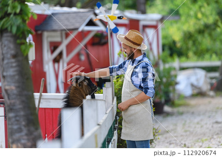 Portrait of farmer petting goat at his farm. Man with hat working on family ranch. 112920674