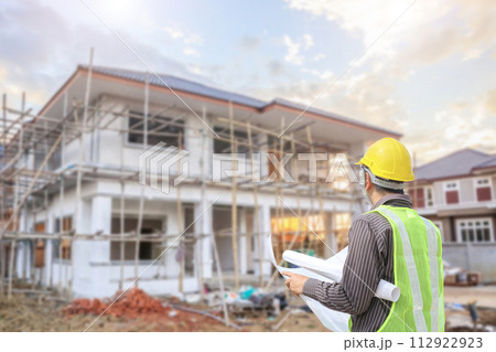 young professional engineer in protective helmet and blueprints paper at the house building construction site 112922923