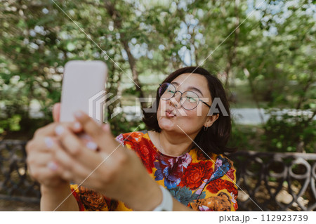 Business casual professional taking a selfie on her cell phone while enjoying nature. Adult asian,entrepreneur with digital devices capturing selfie, staying connected in an urban green space 112923739