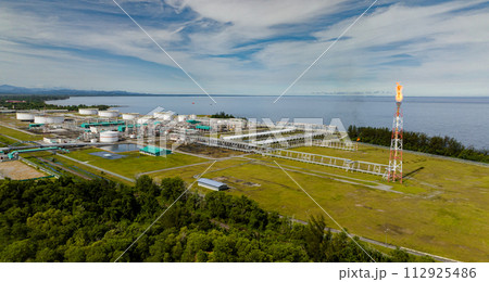 Oil and gas terminal with flaring burning of combustible exhaust gases during petroleum production. Oil storage tanks on the ground in oil distribution station. Borneo, Sabah, Malaysia. 112925486