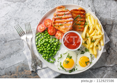 Tender grilled steak served with crisp golden French fries, fied eggs, green pea and fresh tomato closeup on the plate. Horizontal top view Tender grilled steak served with crisp golden French fries, fied eggs, green pea and fresh tomato closeup on the plate. Horizontal top view 112925586