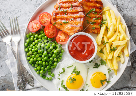 Hearty dinner of grilled loin steak with green peas, fried eggs, french fries, fresh tomatoes and sauce close-up in a plate. Horizontal top view Hearty dinner of grilled loin steak with green peas, fried eggs, french fries, fresh tomatoes and sauce close-up in a plate. Horizontal top view 112925587