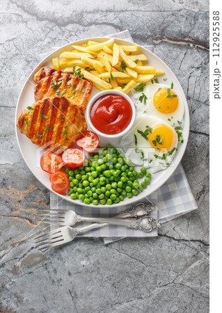 Delicious grilled loin steak with green peas, fried eggs, french fries, tomatoes and sauce close-up in a plate. Vertical top view Delicious grilled loin steak with green peas, fried eggs, french fries, tomatoes and sauce close-up in a plate. Vertical top view 112925588
