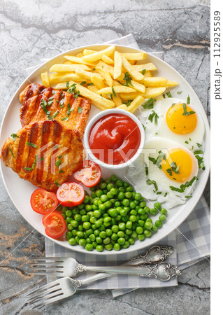 Set lunch Grilled steak with green peas, fried egg, french fries, tomatoes and sauce close-up in a plate. Vertical top view 112925589