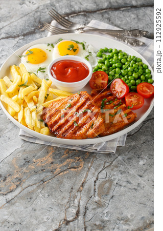 Delicious grilled loin steak with green peas, fried eggs, french fries, tomatoes and sauce close-up in a plate. Vertical 112925592