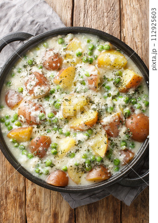Homemade russet potatoes and green peas in creamy cheese sauce close-up in a frying pan on a wooden table. Vertical top view 112925603